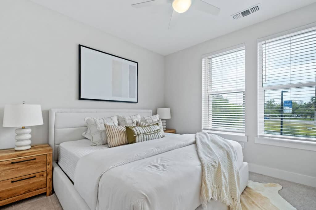 Bright bedroom with a large bed, two wooden nightstands with lamps, white bedding, a blanket, and a framed abstract artwork above the headboard. Two windows let in natural light.