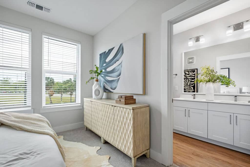 Bright bedroom with a light wood dresser, abstract leaf artwork, and large windows; view into a modern, white bathroom with dual sinks and decorative plants.