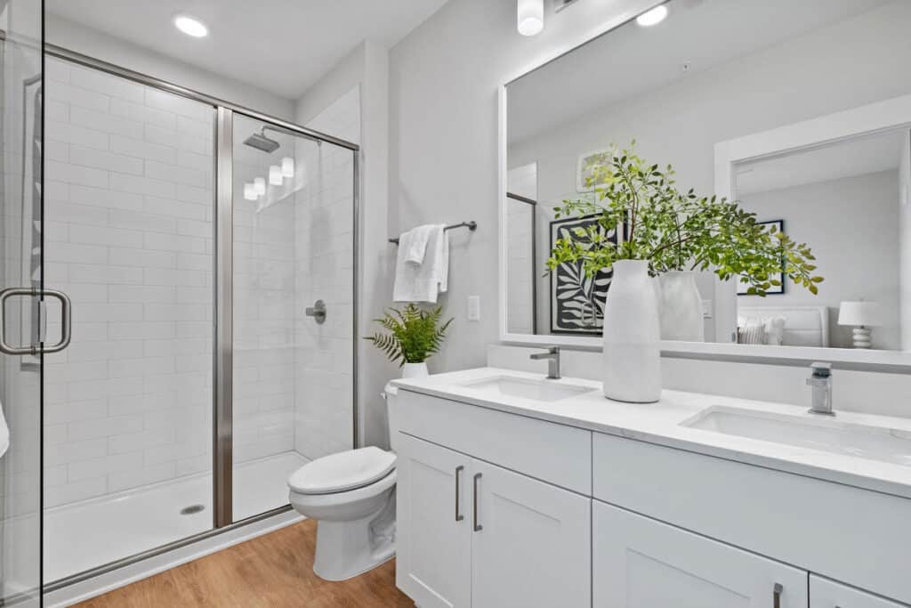 Modern bathroom with glass shower, white vanity with double sinks, a large mirror, potted plants, and wood-look flooring. Towels and soap dispenser are also visible.