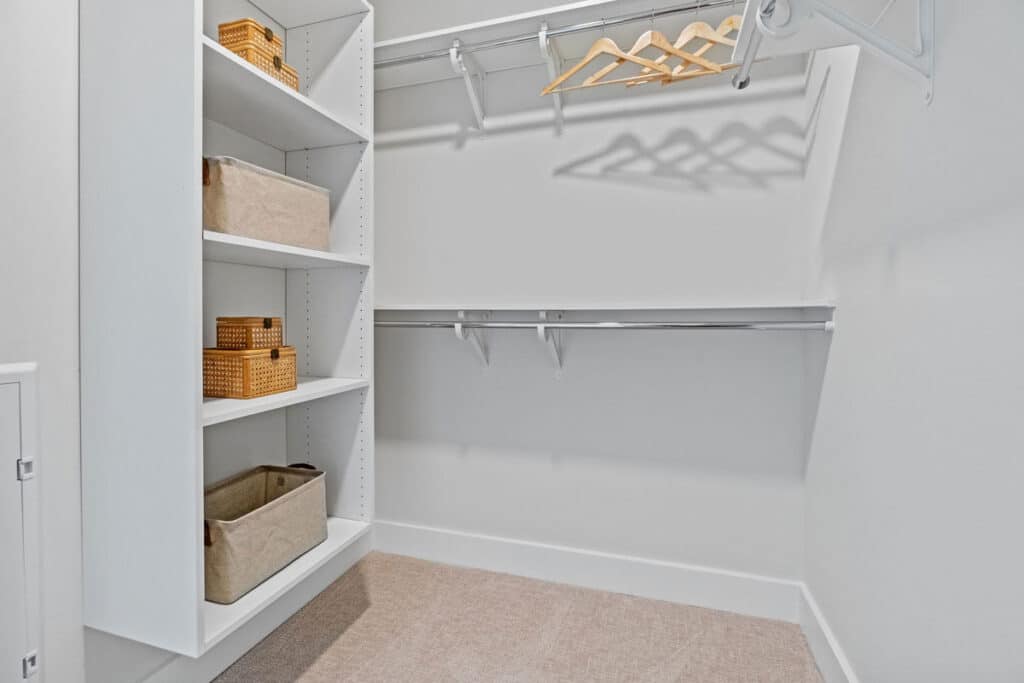 A walk-in closet with white shelves holding baskets and bins, empty hanging rods, and several wooden hangers.