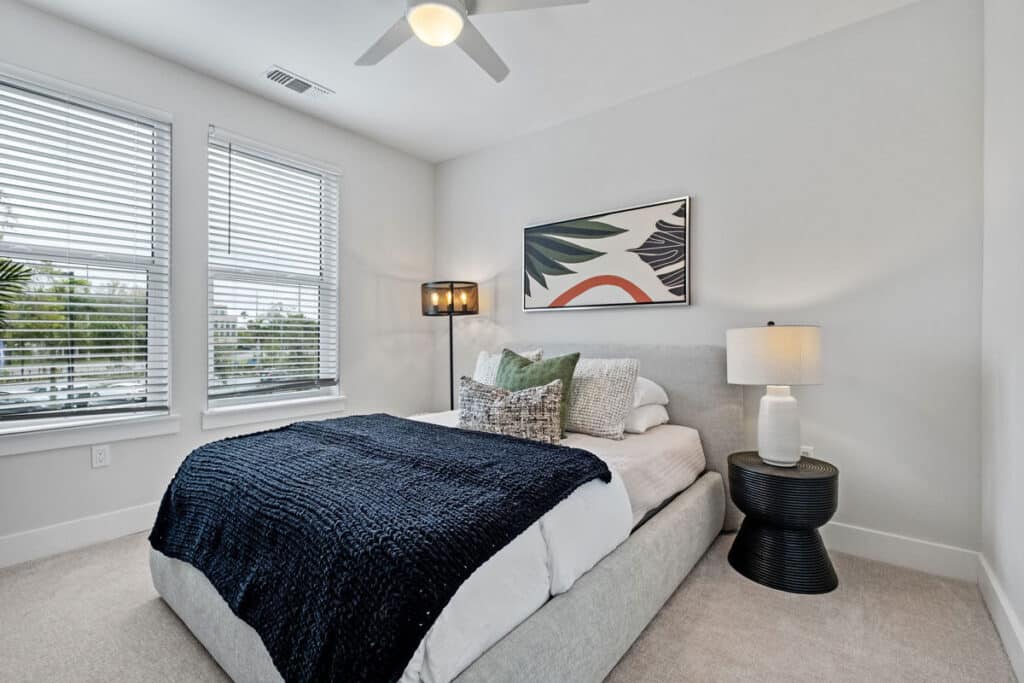 A modern bedroom with a neatly made bed, dark blue blanket, two pillows, a round black nightstand with a lamp, a standing lamp, and a large window with blinds.