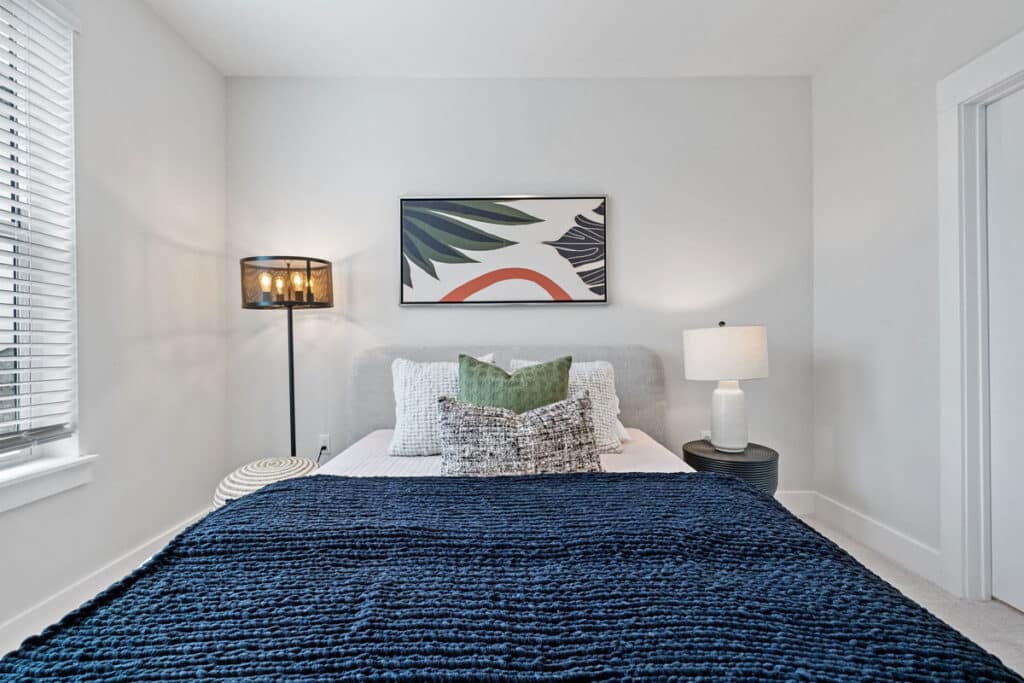 A neatly made bed with a dark blue blanket, decorative pillows, a floor lamp, a table lamp on a nightstand, and abstract art on the wall in a bright, modern bedroom.