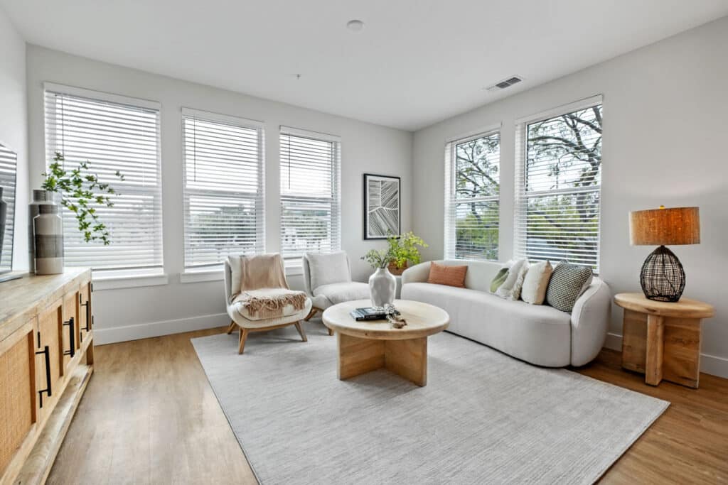 Modern living room with large windows, white walls, light wood furniture, a white sofa, round coffee table, armchair, and decorative accents on a light gray rug.