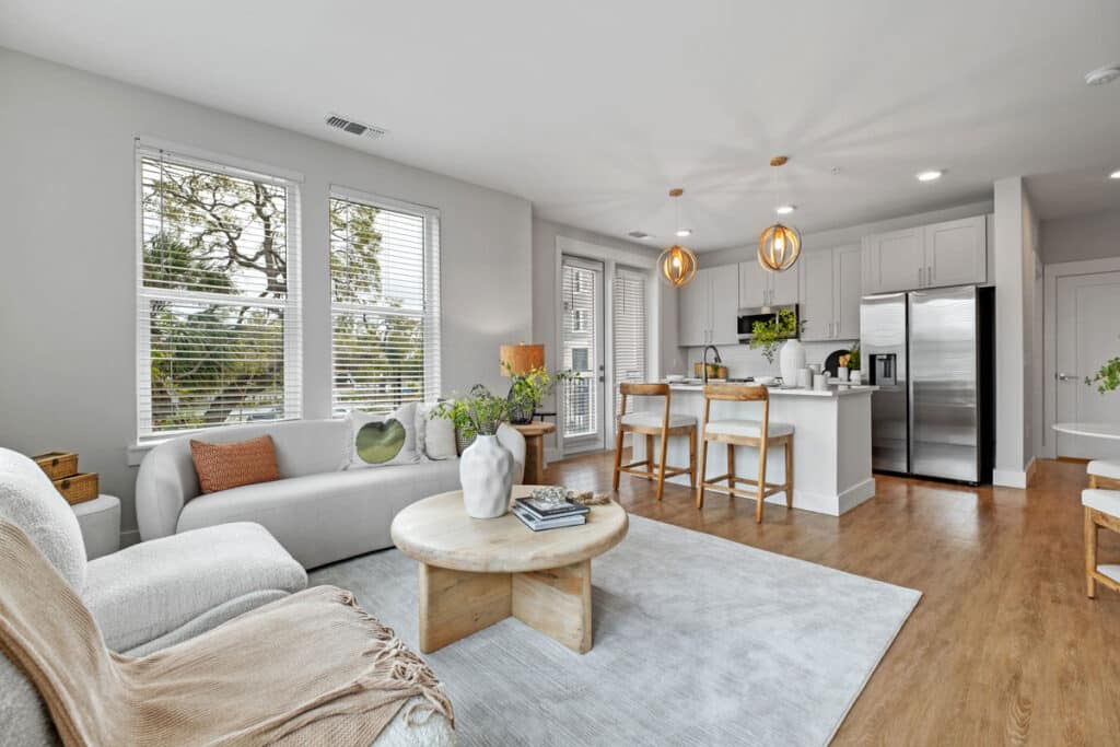 Modern open-concept living room and kitchen with light wood floors, white furniture, large windows, and contemporary decor.