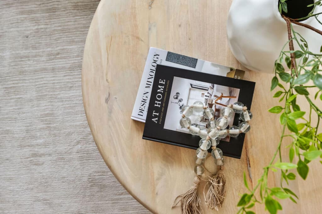 Two stacked design books and decorative beads rest on a light wooden table next to a white vase with green foliage.