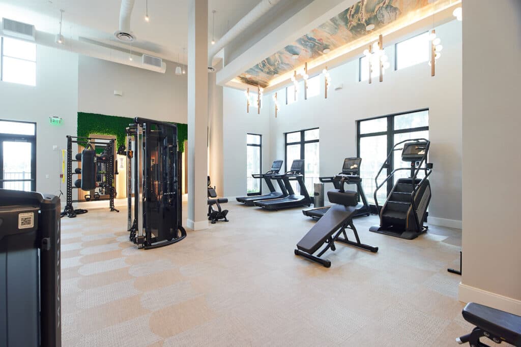 Spacious gym with exercise machines, treadmills, stair climber, adjustable benches, large windows, and modern lighting.