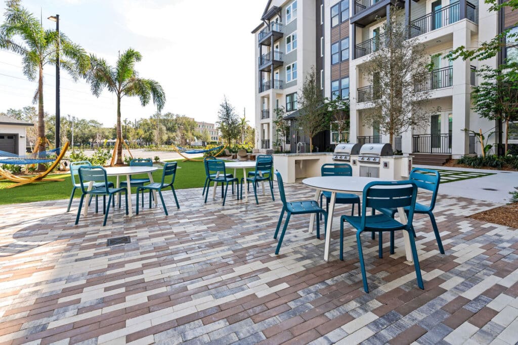 Outdoor patio area with tables, blue chairs, barbecue grills, and hammocks, set beside a modern apartment building and landscaped greenery.