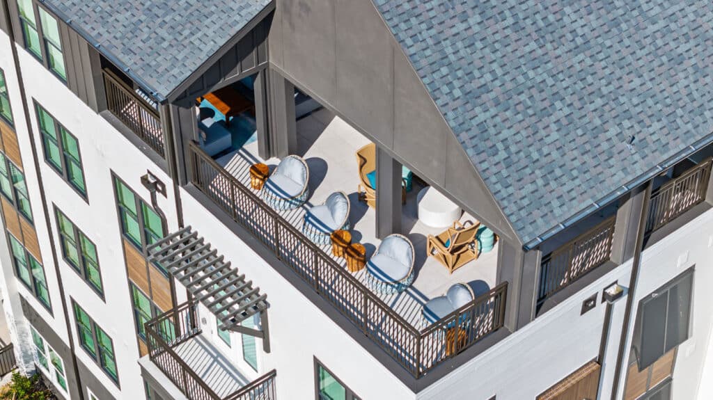 Aerial view of a modern rooftop patio with outdoor seating, tables, and lounge chairs, attached to a white multi-story building with green-trimmed windows.