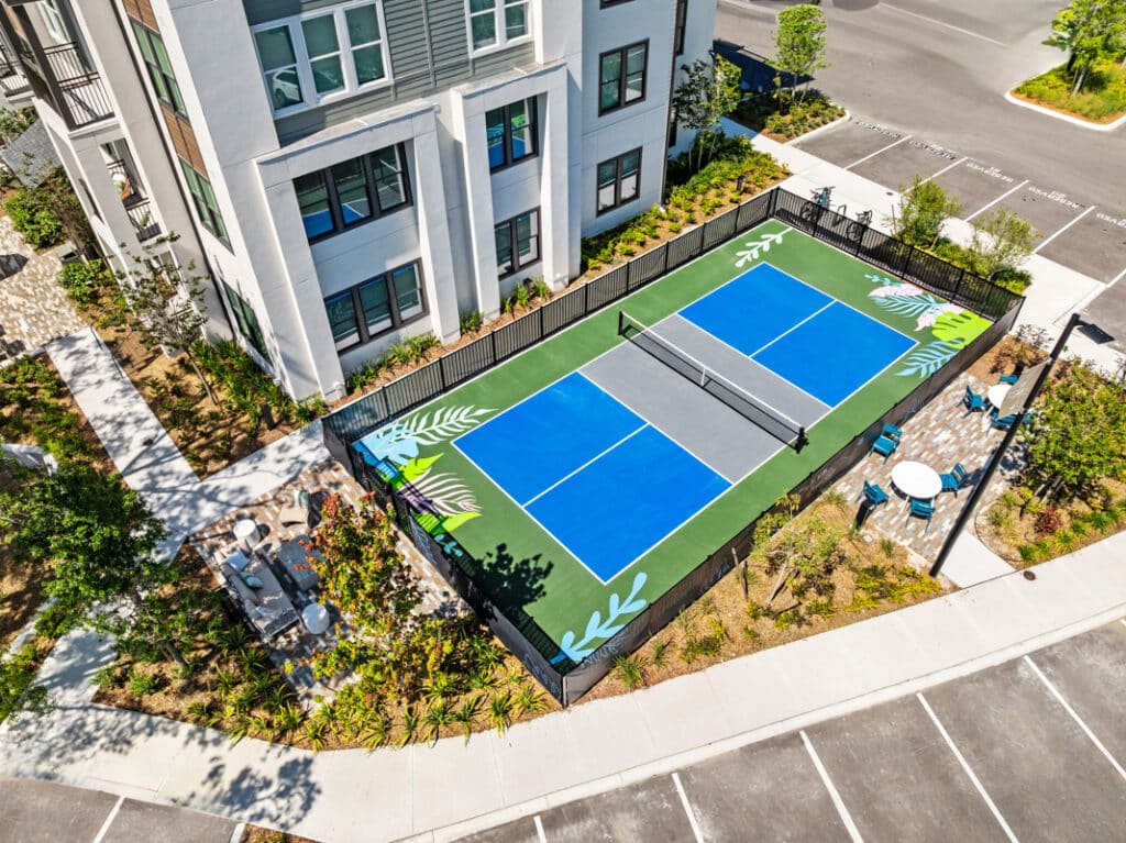 Aerial view of a pickleball court beside a modern apartment building, surrounded by landscaping, pathways, and an adjacent seating area.