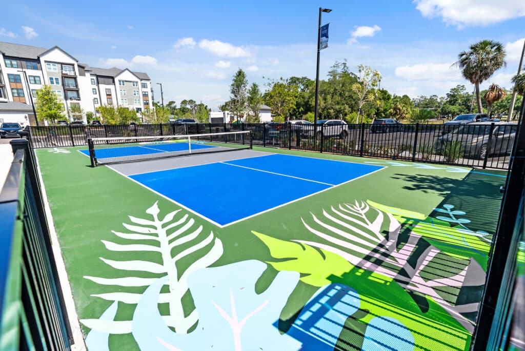 A blue outdoor pickleball court with leaf designs on the surface, surrounded by a black fence, with apartments, parked cars, and trees in the background.