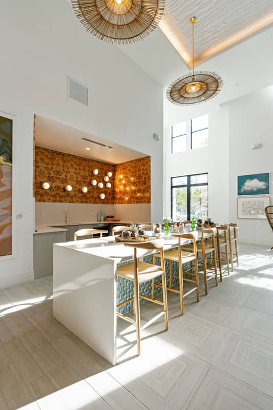 Modern kitchen and dining area with a long island, wooden bar stools, high ceiling, patterned backsplash, pendant lights, and large windows letting in natural light.