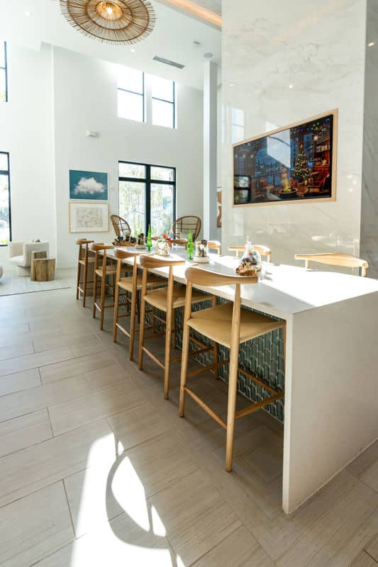 A modern dining area with a long white table, wooden chairs, wall art, large windows, and bright natural light.