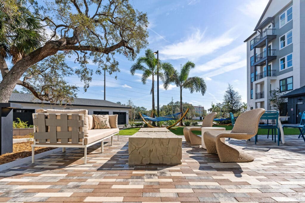 Outdoor patio with modern seating, stone table, and hammocks under palm trees, adjacent to a multi-story residential building on a sunny day.