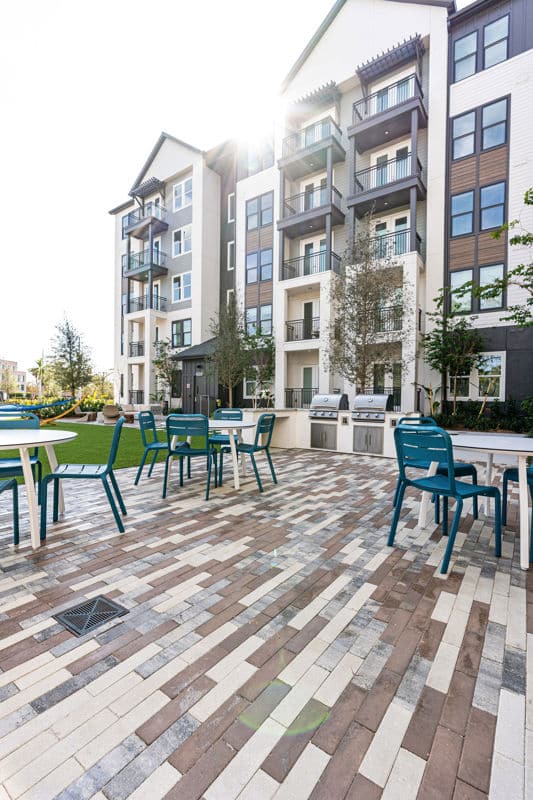 Outdoor patio area with tables, chairs, and barbecue grills in front of a modern multi-story apartment building on a sunny day.