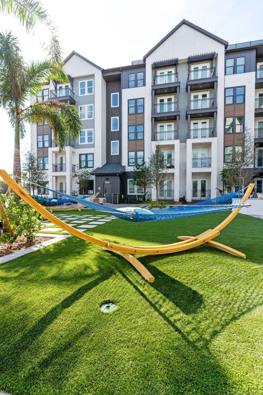 A blue hammock on a wooden stand sits on artificial grass in front of a modern apartment building with balconies and palm trees.