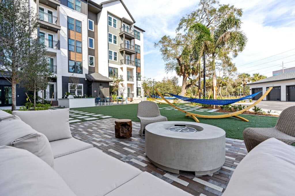 Modern apartment building courtyard with outdoor seating, a circular fire pit, wicker chairs, and two hammocks on a grassy area with trees in the background.