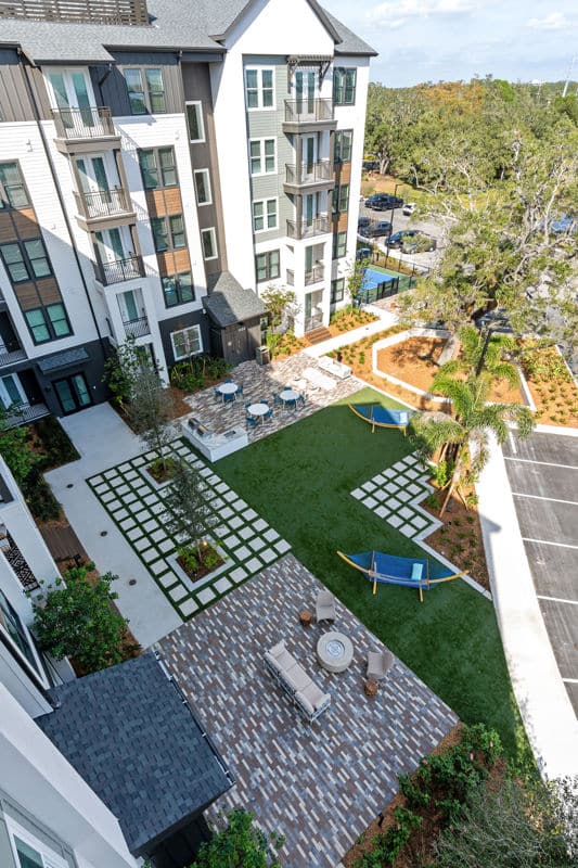 Aerial view of a modern apartment courtyard with green lawn, paved patios, outdoor furniture, and two blue hammocks.