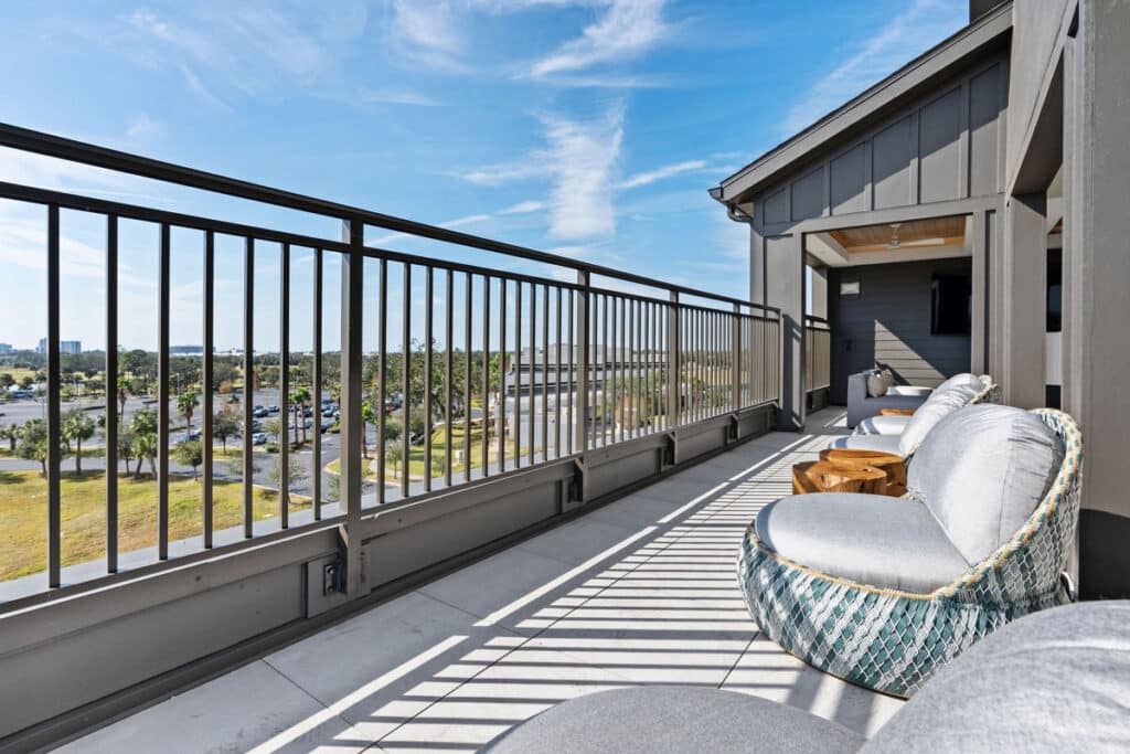 A modern balcony with round cushioned chairs and a small wooden table overlooks a parking lot and trees under a clear blue sky.