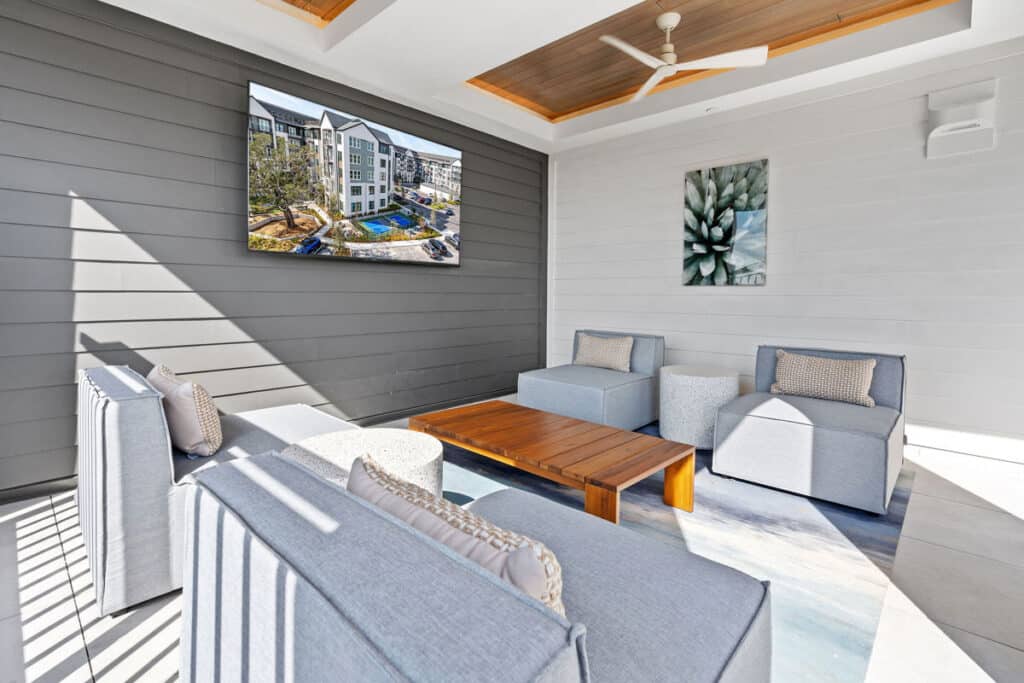 Modern outdoor lounge area with gray cushioned seating, wooden coffee table, wall-mounted TV, ceiling fan, and a flower artwork on the wall. Sunlight enters from the left side.