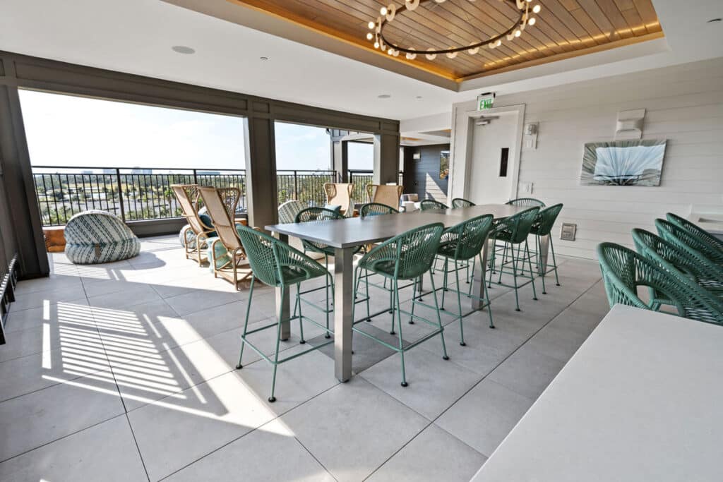 A modern indoor-outdoor lounge with green chairs, long tables, wicker seating, large windows, and a view of the outdoors.