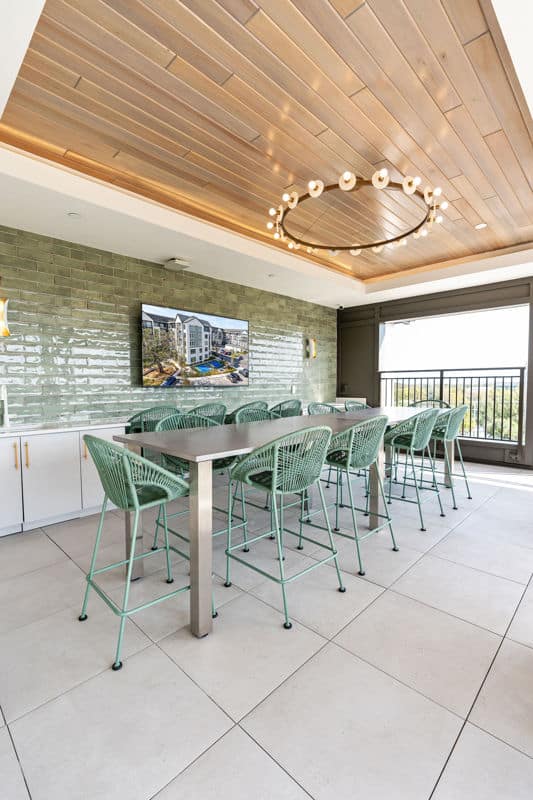 Modern outdoor dining area with a long table, mint green chairs, a wall-mounted TV, green tile wall, and a circular ceiling light fixture.