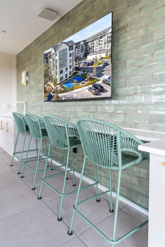 Four green bar stools are lined up at a white counter against a green tiled wall, with a large screen displaying an apartment complex above them.