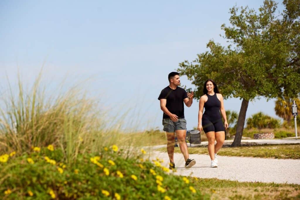 Two people in athletic clothing walk and talk on a gravel path in a park with trees and yellow flowers on a sunny day.