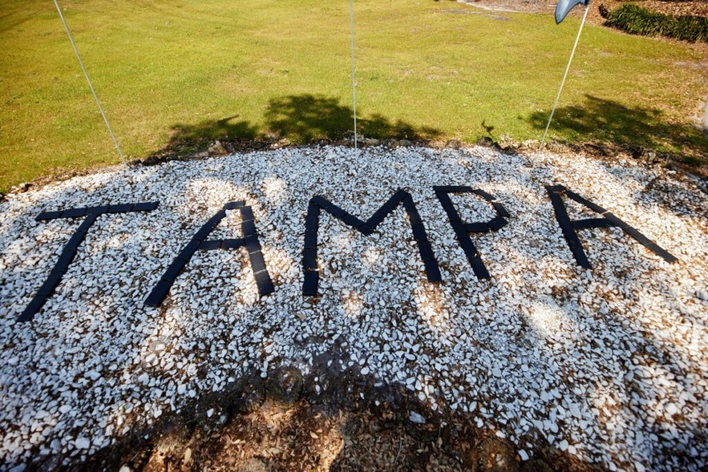 The word "TAMPA" spelled out in large black letters on a bed of white rocks, with green grass in the background.