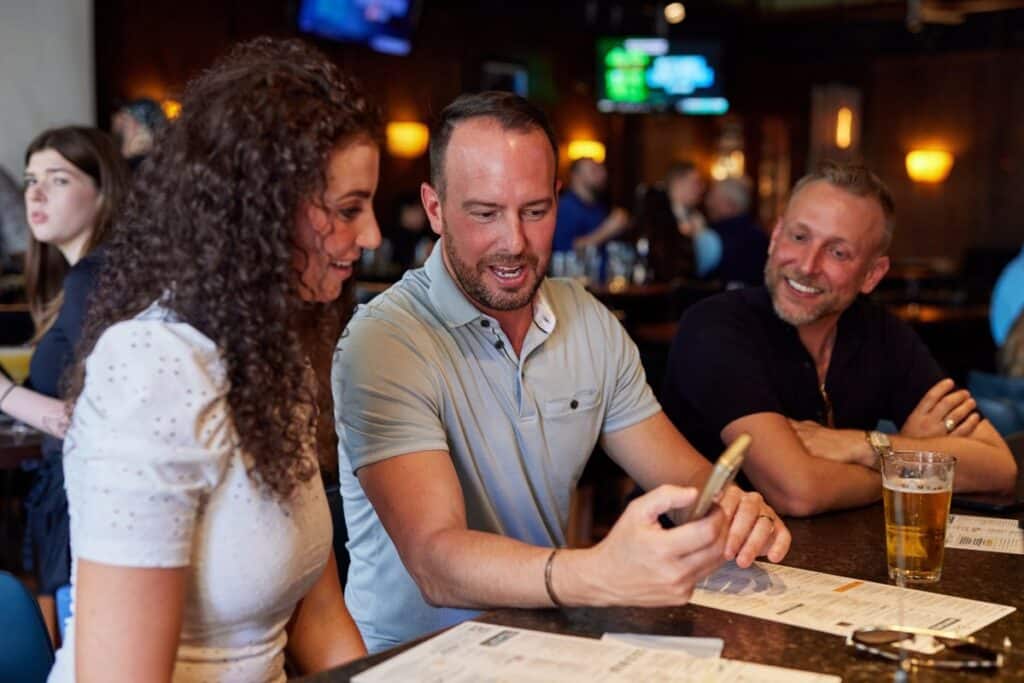 Three people sit at a bar, looking at a phone and smiling. Menus and drinks are on the counter in front of them. Other patrons are visible in the background.