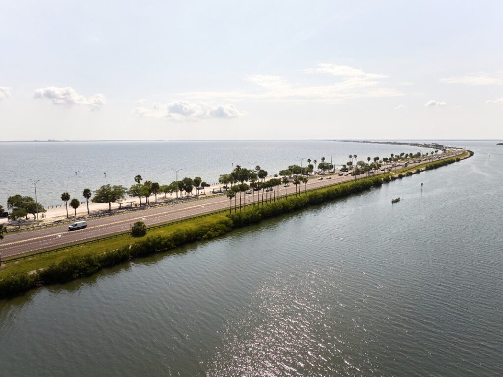 A narrow road lined with palm trees runs between a body of water and a bay, with cars driving in both directions under a partly cloudy sky.