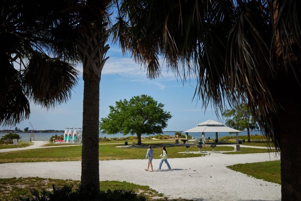 Two people walk on a gravel path in a sunny park with palm trees, open shelters, and a view of the water in the background.