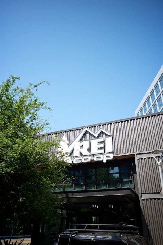 The exterior of an REI Co-op store with a large sign on the building, trees nearby, and a clear blue sky overhead.