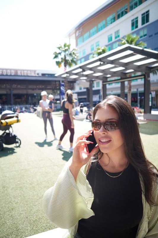 A woman wearing sunglasses talks on her phone in an outdoor plaza, with people walking and a restaurant in the background on a sunny day.