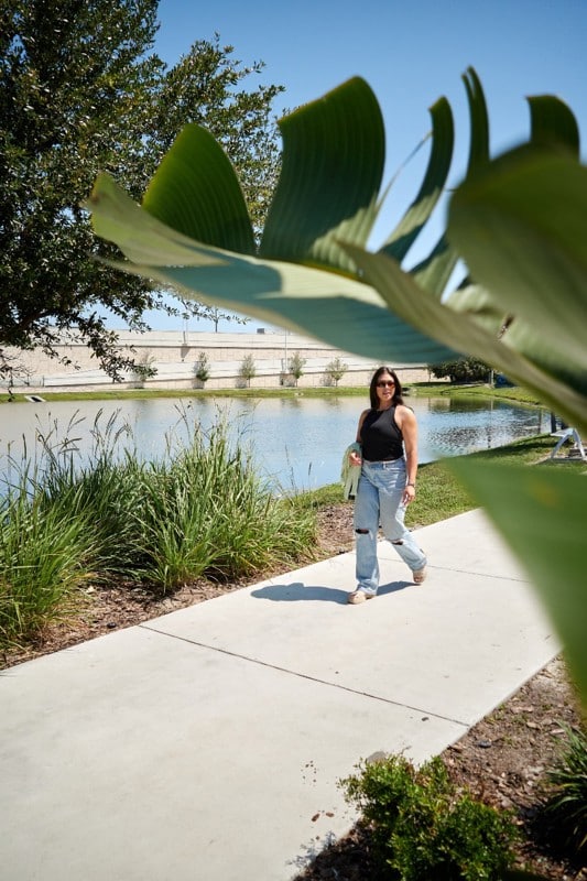A woman in sunglasses walks on a sidewalk beside a pond, partially framed by large green leaves in the foreground.