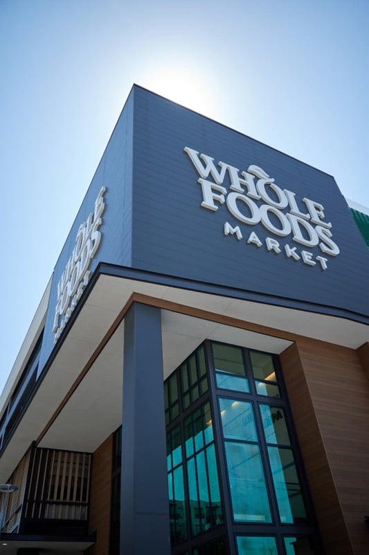 The exterior of a Whole Foods Market store with large signage, modern glass windows, and the sun visible behind the building.