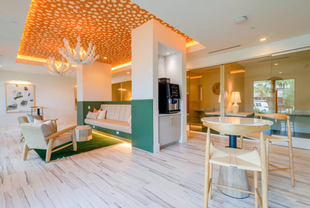 Modern lounge area with patterned orange ceiling, green cushioned bench, beverage machine, light wood furniture, and large glass doors.