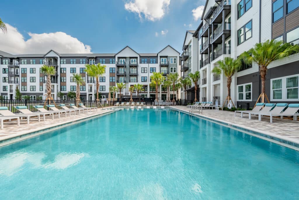 Rectangular outdoor swimming pool surrounded by lounge chairs, palm trees, and modern multi-story apartment buildings under a partly cloudy sky.