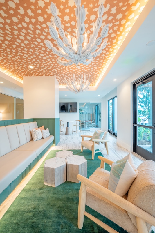Modern lounge area with a green rug, light wood furniture, geometric coffee tables, large windows, and a patterned ceiling with white decorative light fixtures.