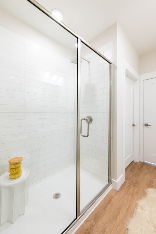 A modern bathroom with a glass-enclosed shower, white subway tiles, a small white stool with sponges, and light wood flooring.