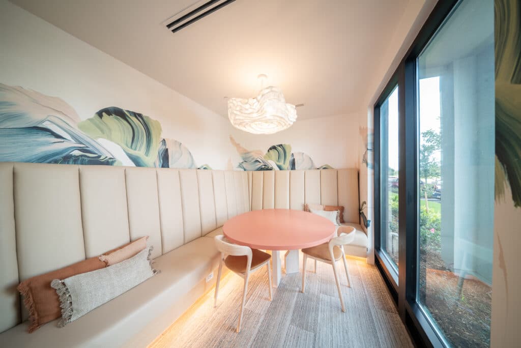 A small, modern meeting room features a round pink table, four chairs, beige cushioned benches, wall art with large leaves, and a large window letting in natural light.