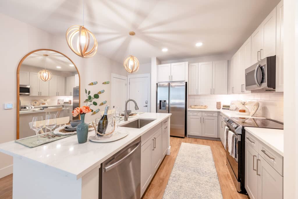 Modern kitchen with white cabinets, stainless steel appliances, an island with a sink, pendant lights, a large round mirror, and decorative items arranged on counters and the island.