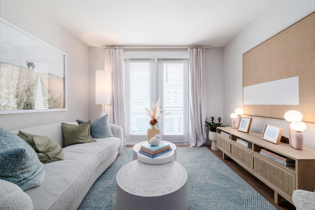 A bright, modern living room with a white sofa, green pillows, a round coffee table, light wood console, abstract art, and large windows with sheer curtains.