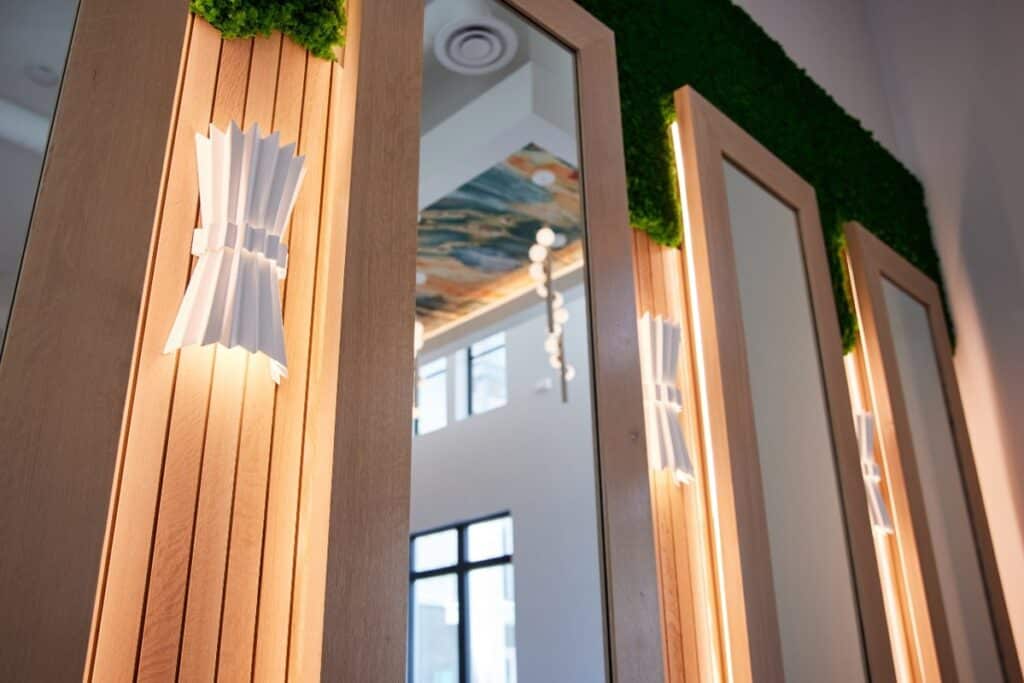 Vertical wooden wall panels with built-in lights, white sconces, and mirrors, decorated with artificial green foliage at the top, in a modern interior space.