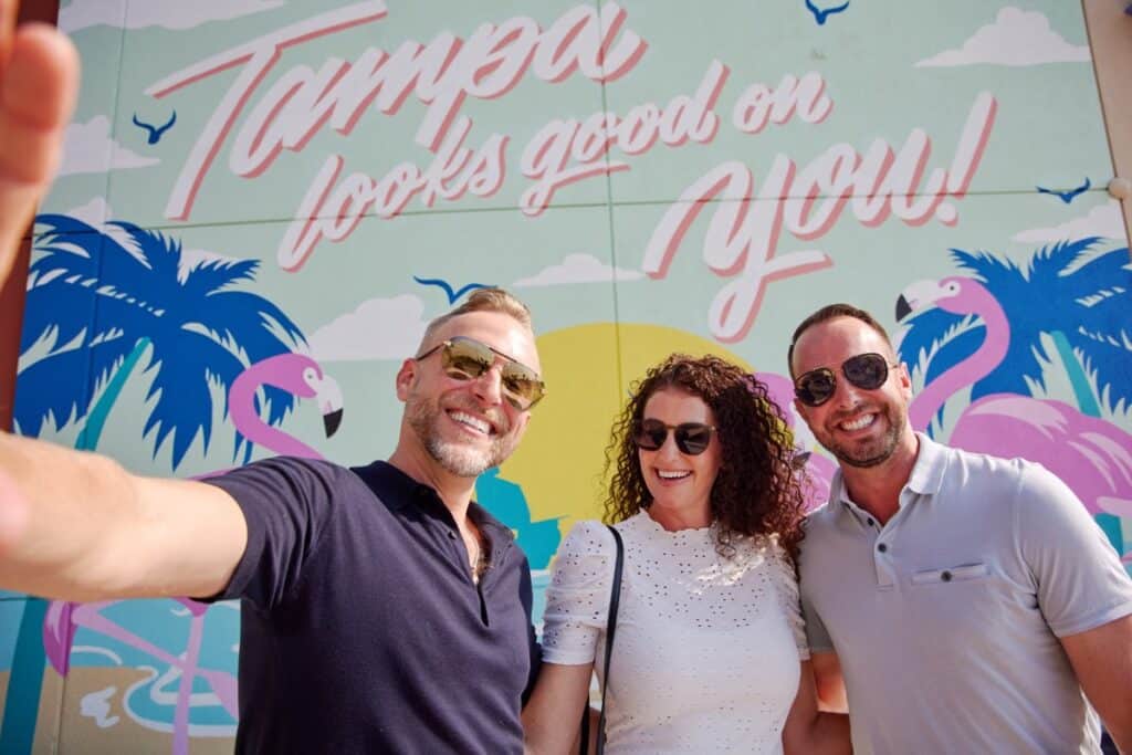 Three people smiling and taking a selfie in front of a mural that reads "Tampa looks good on you!" with flamingos, palm trees, and a sun in the background.