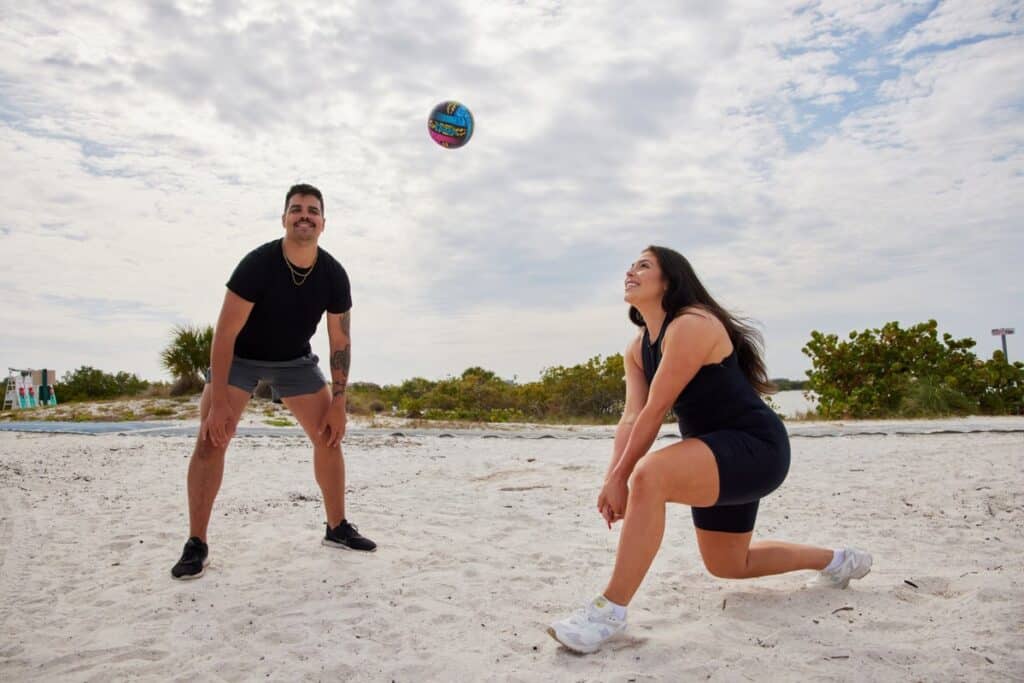 Two people play beach volleyball on white sand under cloudy skies; one stands ready while the other crouches to receive the ball.