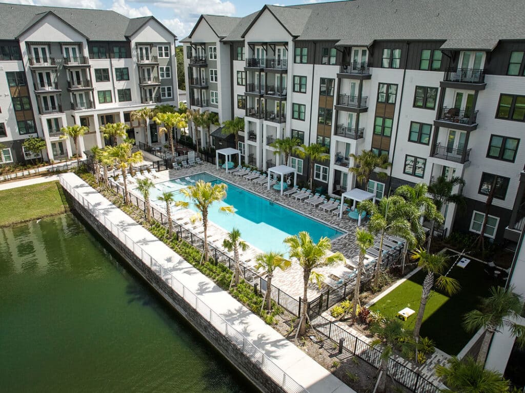 A large outdoor swimming pool with lounge chairs and cabanas, surrounded by palm trees, is situated in the courtyard of a modern apartment complex next to a pond.