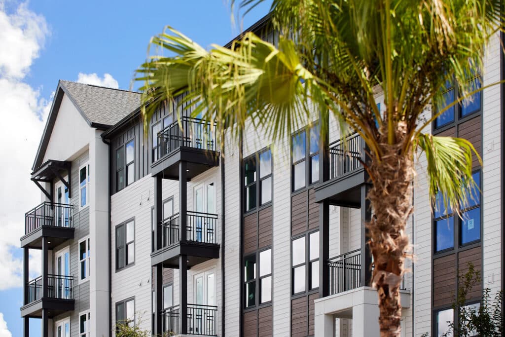 Modern apartment building with multiple balconies and large windows, partially obscured by a tall palm tree under a blue sky with clouds.