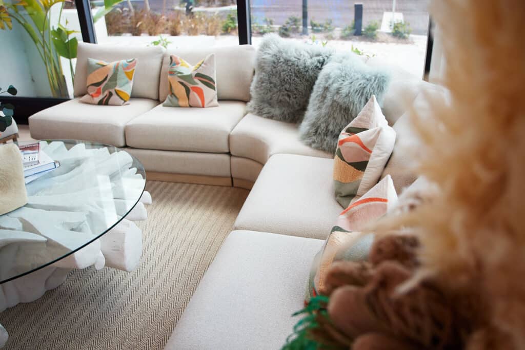 A modern living room features a curved white sectional sofa with patterned and furry pillows, a round glass coffee table, and large windows.
