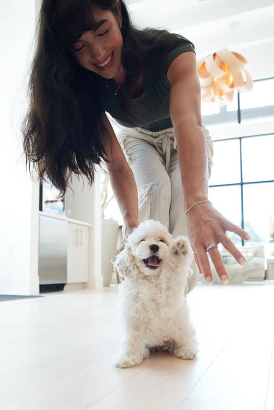 A woman smiles and leans down toward a small, fluffy white puppy standing on a light-colored floor with its paw raised.