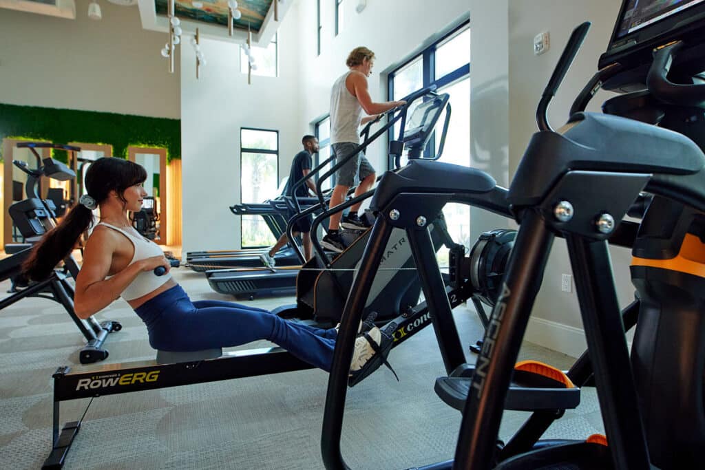 Three people exercising in a gym: one woman using a rowing machine and two others using stair climber machines. Fitness equipment is visible in a brightly lit room.
